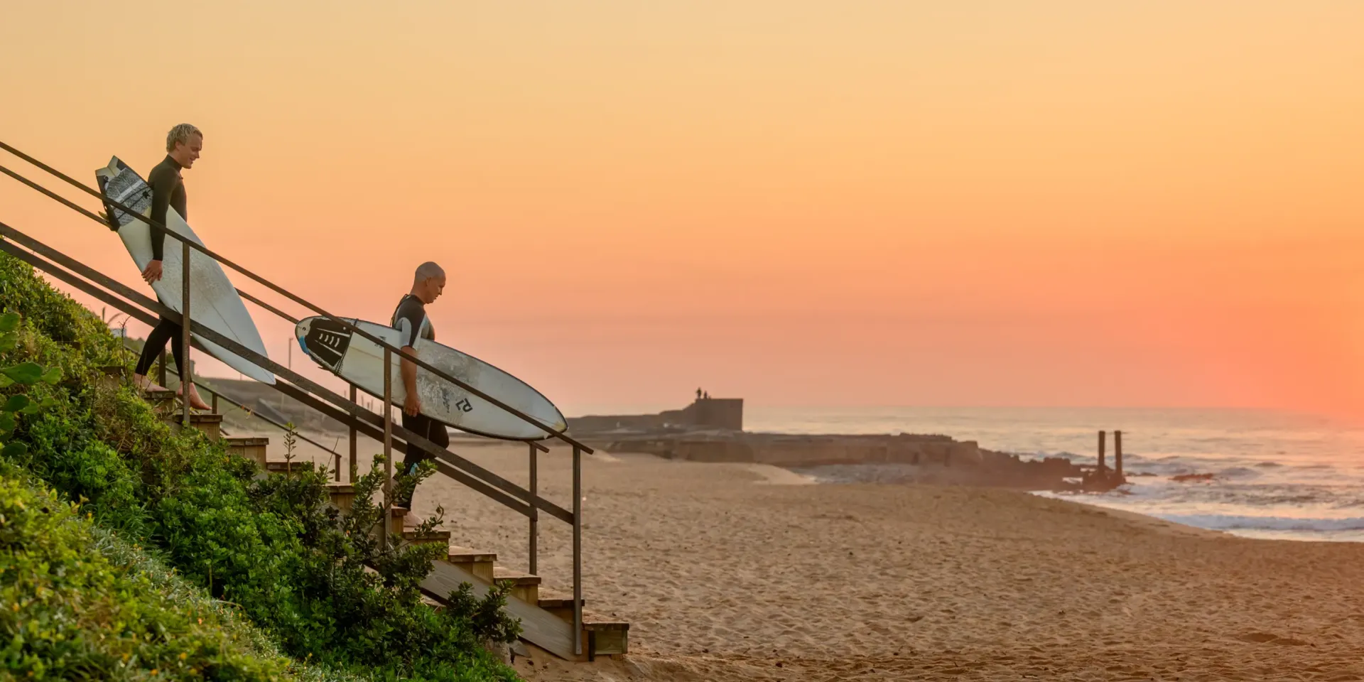 Two surfers carrying their boards down the steps to Salt Rock beach at sunset